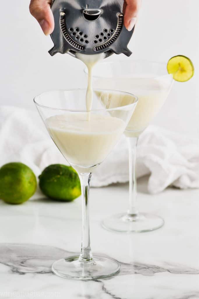 cocktail strainer pouring a white key lime martini recipe into a martini glass, another glass in the background that is full and garnished with a key lime wedge