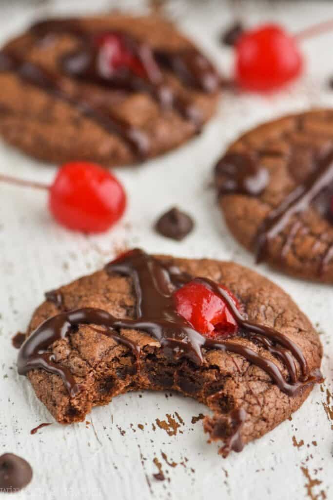 a fudgey chocolate covered cherry cookie that has had a bite taken out of it sitting on a white wood board with more cookies in the background