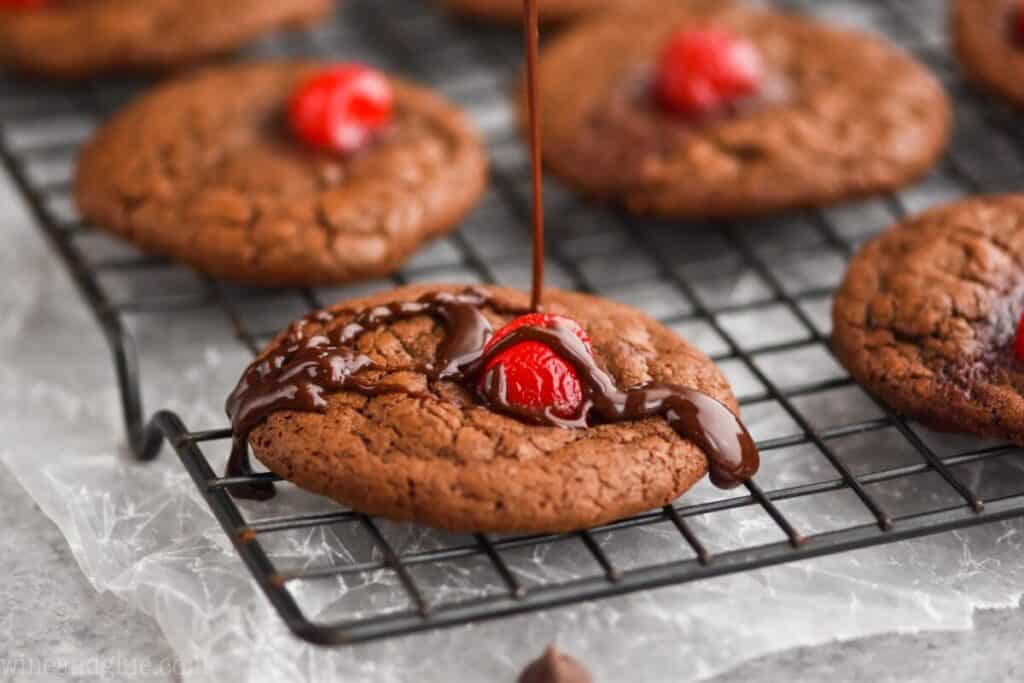 a landscape photo of a chocolate covered cherry cookie on a cooling wrack that is being drizzled with chocolate ganache