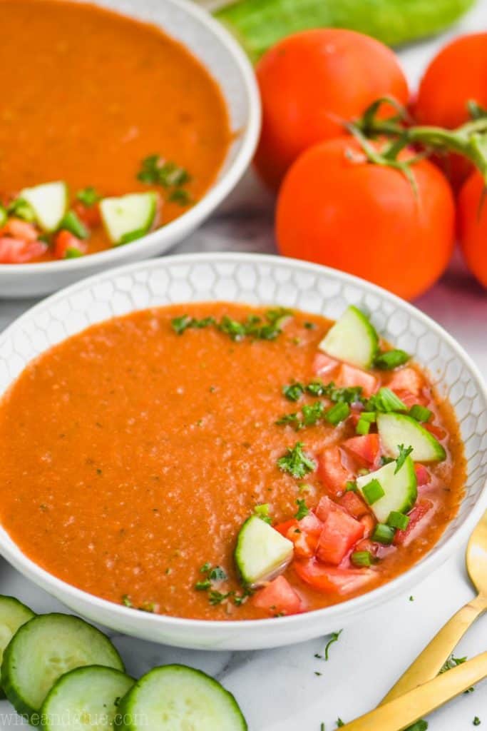 white bowl full of easy gazpacho recipe topped with chopped tomatoes cucumbers and parsley, cucumber slices next to the bowl and whole tomatoes behind it