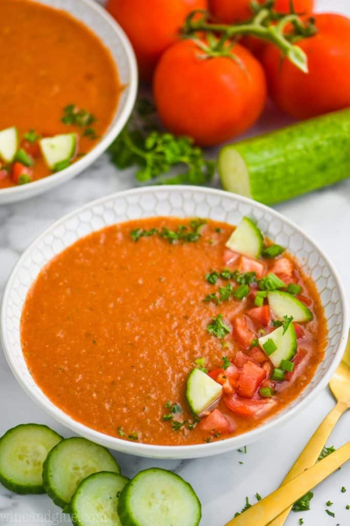 white bowl full of easy gazpacho recipe topped with chopped tomatoes cucumbers and parsley, cucumber slices next to the bowl and whole tomatoes behind it