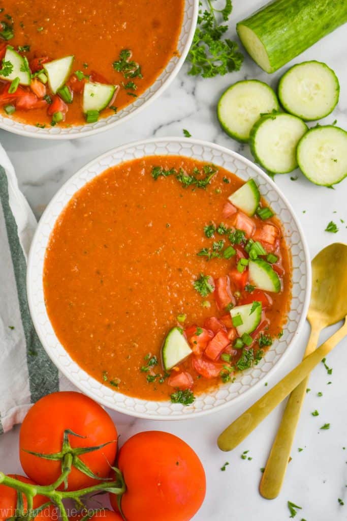 overhead view of gazpacho soup in a white bowl, cut up cucumber and whole tomatoes next to the soup