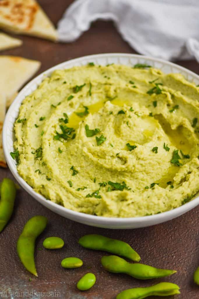 side view of edamame hummus recipe in a white bowl, garnished with parsley and oil with soy beans around the bowl