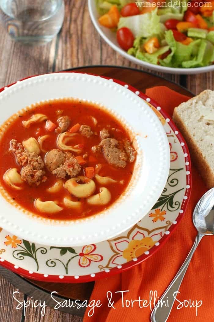 A bowl of the Spicy Sausage and Tortellini Soup with a side of a salad and a bread slice
