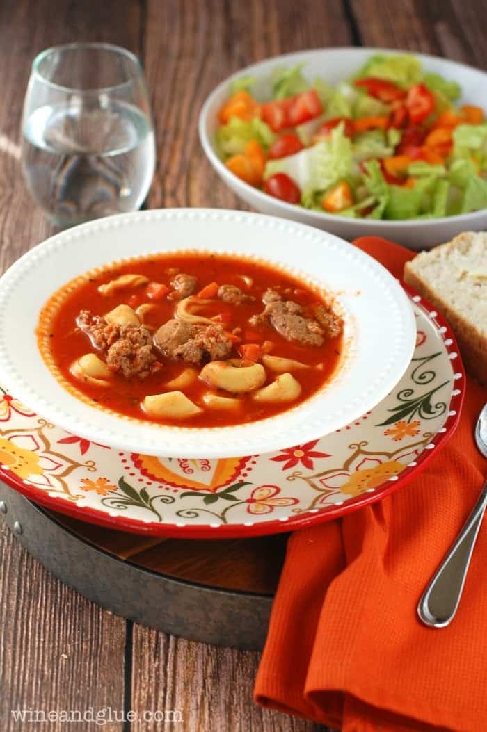 A bowl of the Spicy Sausage and Tortellini Soup with a side of a salad and a bread slice
