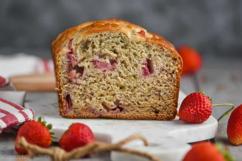 landscape photo of a loaf of strawberry banana bread cut in half, fresh strawberries visible in the bread, and fresh strawberries on the marble cutting board bread is sitting on