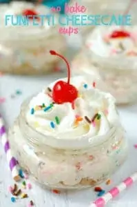 close up of a mason jar with no bake cheesecake, topped with a cherry, whipped cream and sprinkles, says no bake funfetti cheesecake cups