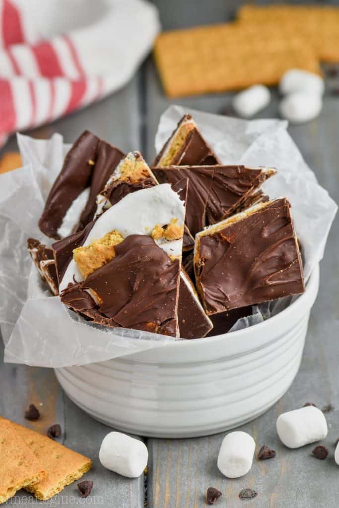 s'mores chocolate bark sticking out of a small white bowl with wax paper, surrounded by graham crackers, mini marshmallows, and mini chocolate chips