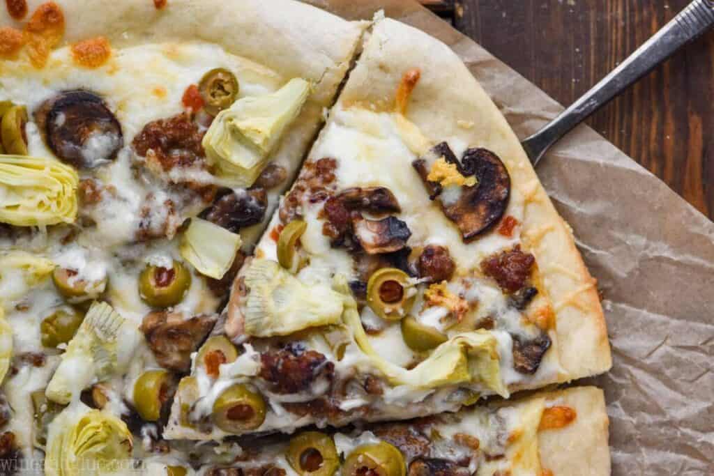 landscape photo of a white pizza topped with olives, artichokes, sausage, and mushrooms, one piece coming out