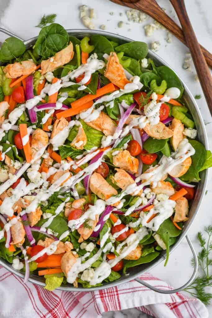 overhead view of a buffalo chicken salad dressed with ranch dressing in a large metal tray with blue cheese, wooden serving utensils, fresh dill, and a red and white napkin around it