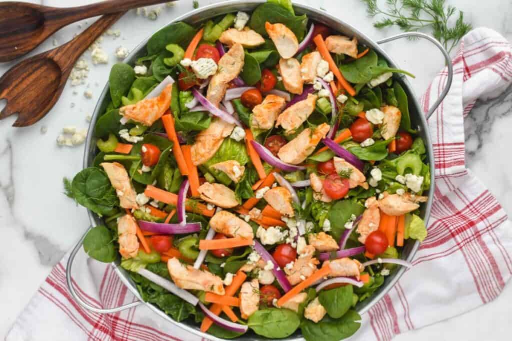 overhead of a buffalo chicken salad recipe in a galvanized metal try with buffalo chicken, spinach, romaine lettuce, carrots, red onions, and tomatoes