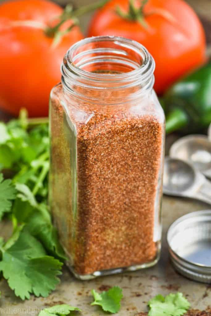 bottle of spice jar with homemade taco seasoning next to cilantro with tomatoes in the background