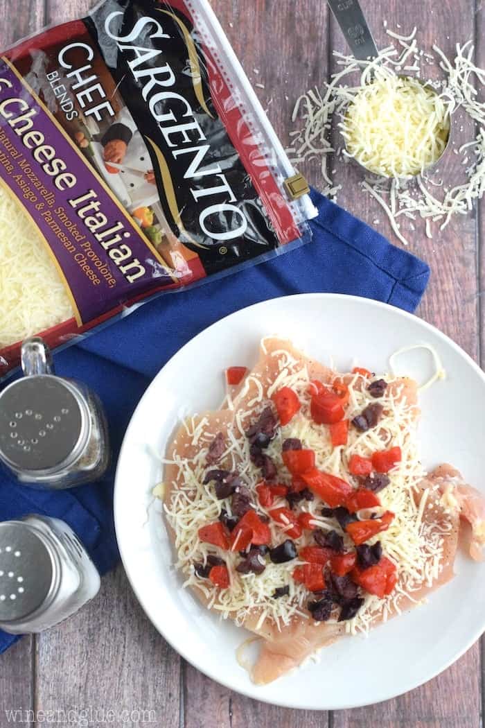 Bird's eye view of a plate that has raw chicken topped with cheese, olives, and chopped pepper. Above the plate is a Sargento's 6 Cheese Italian bag and a measuring cup with some cheese inside.  