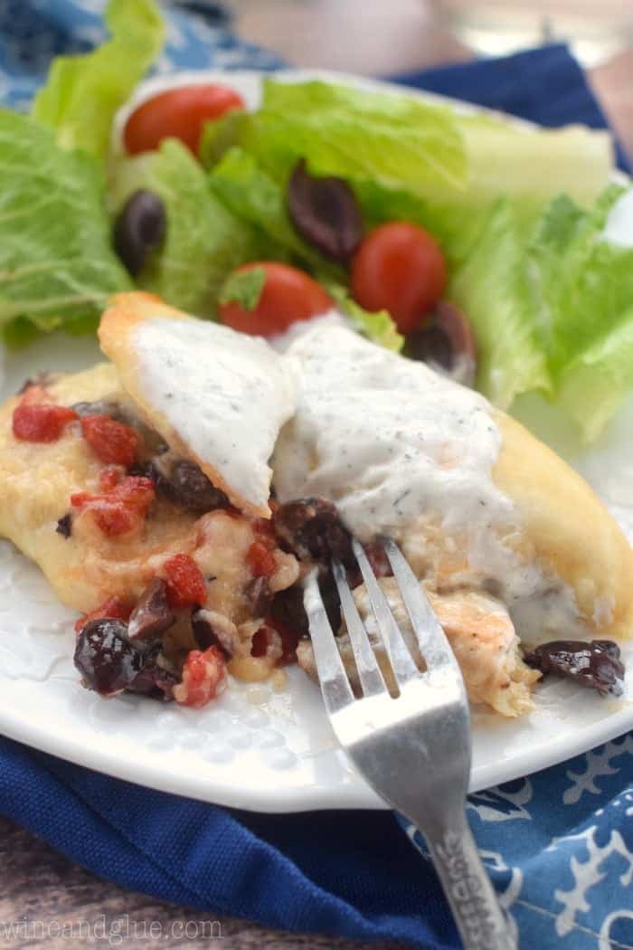 A fork cut into the Greek Stuffed chicken on one side of the plate. On the other is a salad with olives, cherry tomatoes, and lettuce.