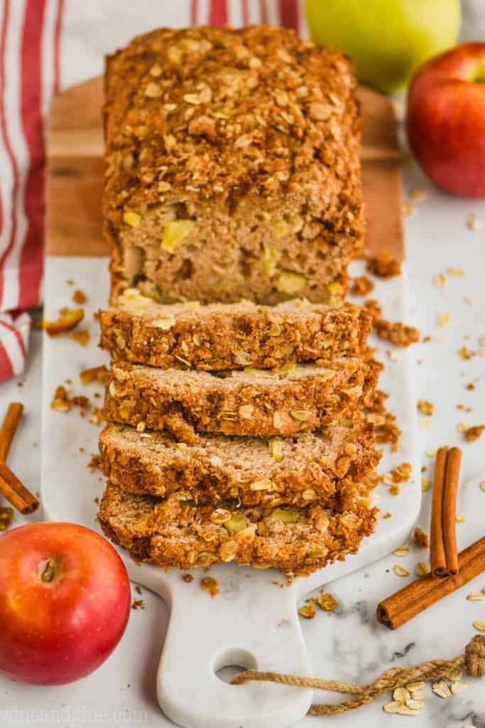 pulled back view of apple bread recipe that has been sliced into on a marble and wood cutting board with oats around, cinnamon sticks, and apples