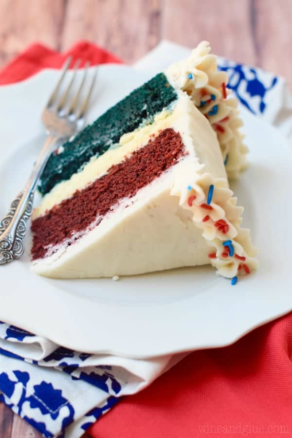 A slice of the Red White and Blue Velvet Cheesecake Cake shows the distinct layers of red, white, and blue and frosted with white frosting. On the top and the bottom edge of the cake, some frosting was piped with a star tip and topped with red, white, and blue sprinkles. 