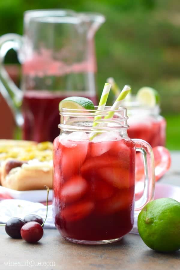 a glass of cherry limeade, garnished with a lime wedge, with a pitcher of it in the background