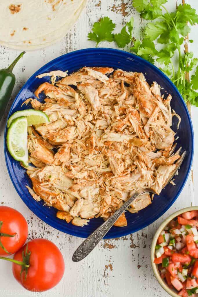 overhead view of shredded chicken for tacos on a blue plate with a lime wedge, cilantro, pico de Gallo, tomatoes, and a jalapeño around the plate