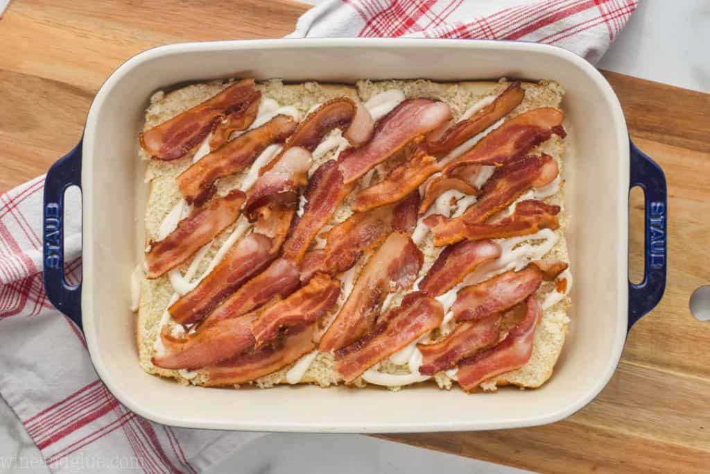 landscape photo of a baking dish on a cutting board with the bottom half of slider rolls, topped with cooked bacon