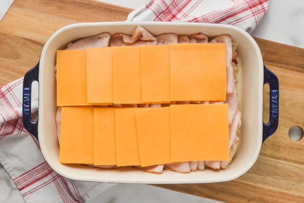 landscape photo of a baking dish on a cutting board with the beginnings of turkey sliders, at the step of adding sliced cheddar cheese