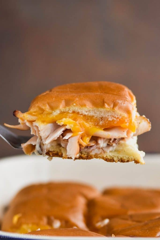 a bacon ranch turkey slider being pulled out of a baking dish of other sliders with melted cheddar cheese and turkey visible