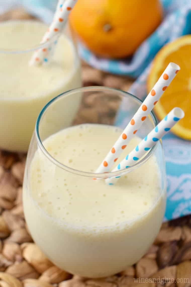 In a glass, the Orange Colada Smoothie has a pastel yellow color and two paper straws with polka dots. 