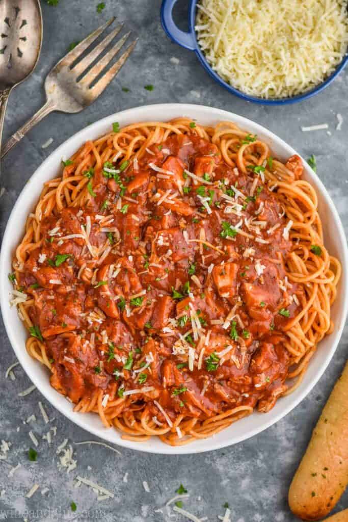 an overhead view of a white bowl full of spaghetti with red sauce on a gray background with a bowl of parmesan cheese and breadsticks next to it