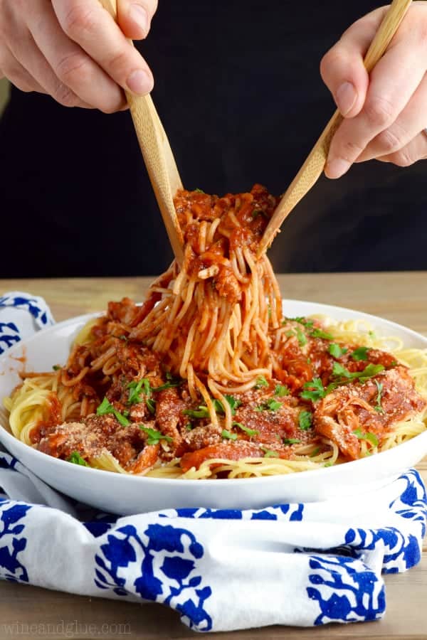 two hands tossing chicken spaghetti in a white serving bowl
