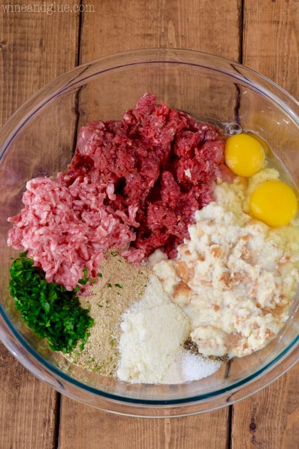 overhead view of a clear glass bowl filled with meat, eggs, soaked bread, fresh parsley, and breadcrumbs to make Italian meatballs