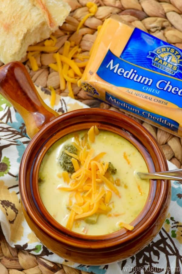 an overhead view of a brown soup bowl filled with crock pot broccoli cheddar soup with a block of cheddar cheese next to it