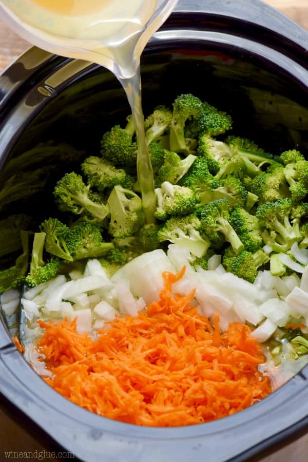 a crock pot filled with shredded carrots, diced onions, and broccoli florets with chicken stock being poured on them for slow cooker broccoli cheddar soup