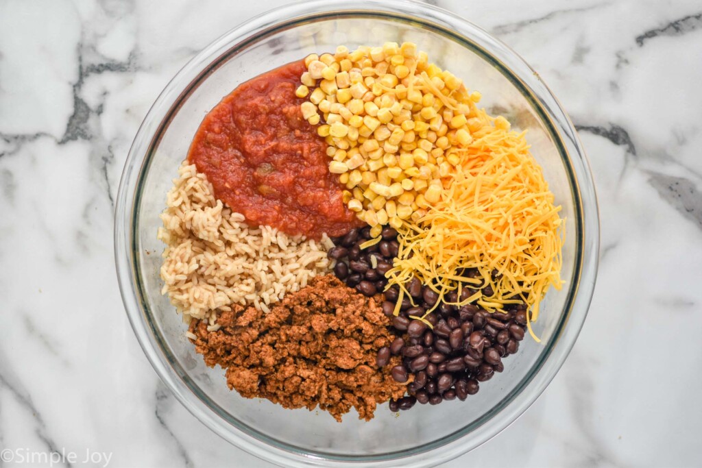 overhead of salsa, corn, cheese, black beans, ground beef with taco seasoning, and rice in a large glass mixing bowl before being mixed together