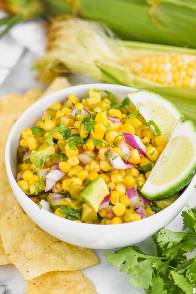 small white bowl with the best corn salsa recipe garnished with two lime wedges, corn on the cob in the background