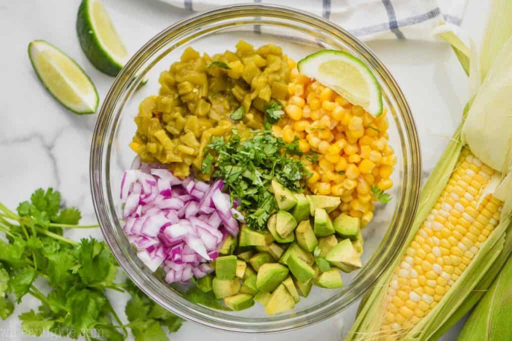 corn salsa ingredients separated by ingredient in a clear bowl with lime wedges, cilantro, and corn on the cob on the side