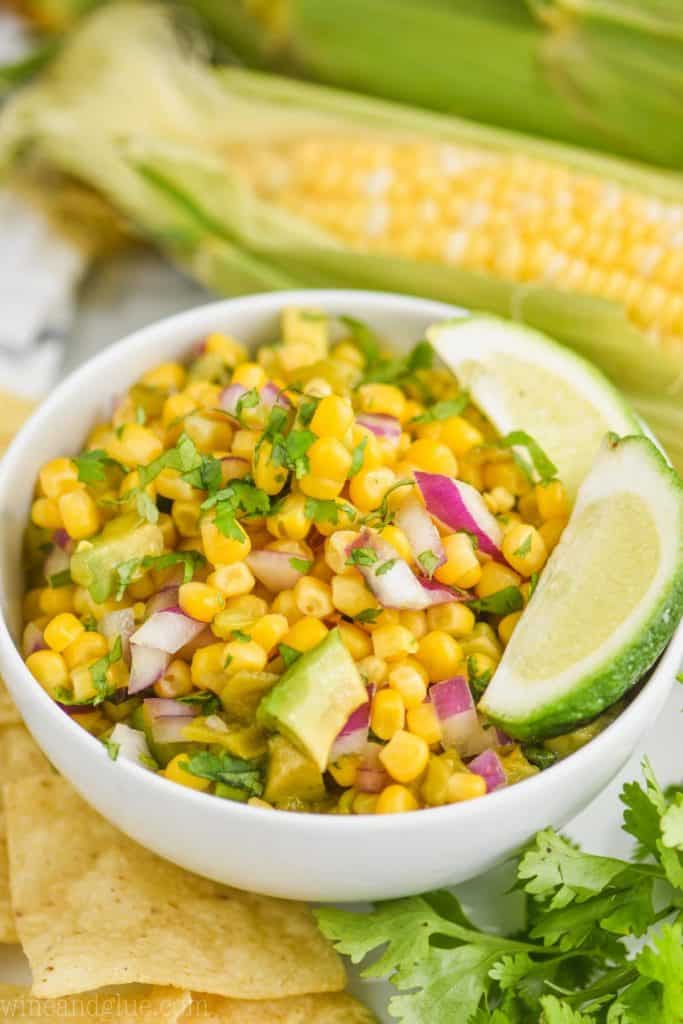 small white bowl filled with corn salsa recipe and garnished with lime wedges, whole corn on the cob in the background