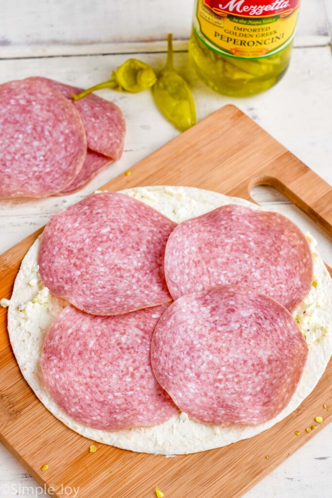 tortilla on a cutting board with cream cheese mixture and four pieces of salami placed evenly on it
