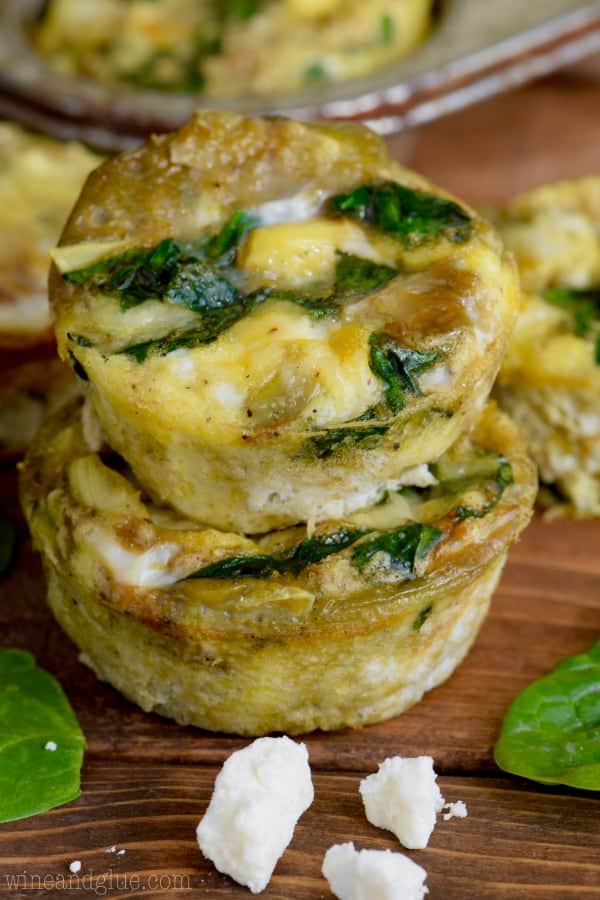 Two Spinach and Artichoke Egg Muffins are stacked on top of each other showing the spinach, melted feta cheese, white onion, and artichoke.