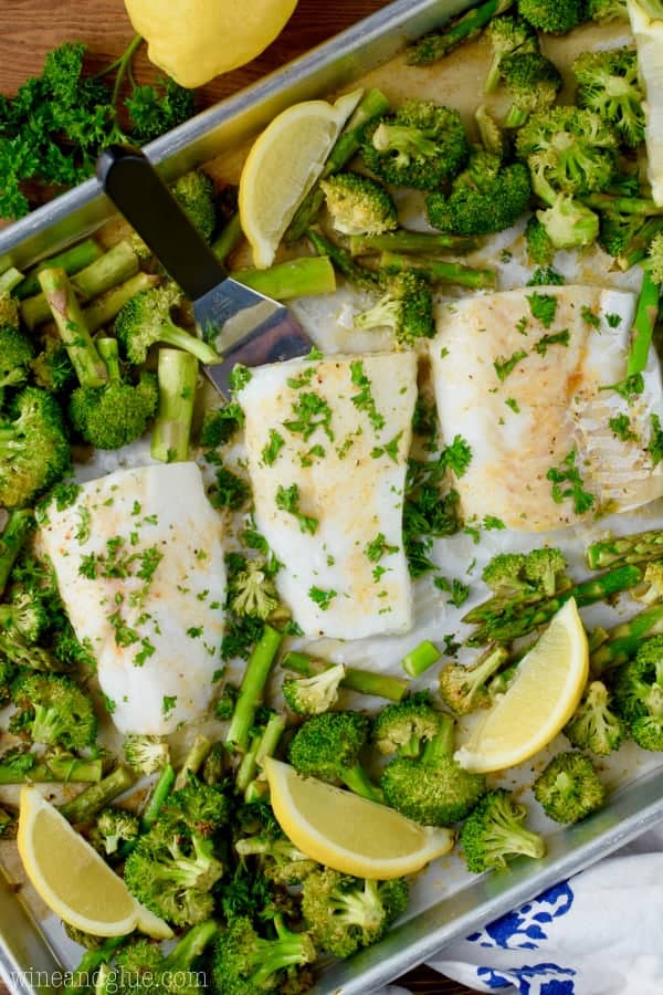 On a sheet pan, three baked cods have a golden crust surrounded by asparagus and broccoli. 