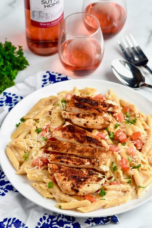 side view of a plate of creamy cajun chicken pasta, an easy chicken recipe garnished with fresh parmesan, parsley and diced tomatoes with two glasses of wine in the background