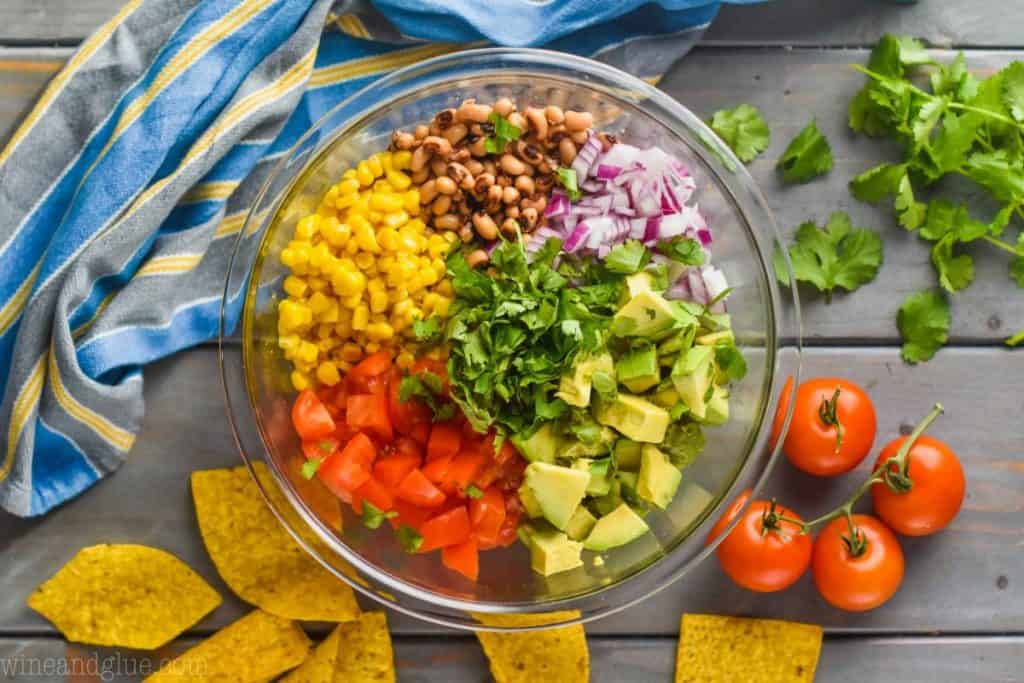 overhead view of a clear bowl that is full of the ingredients for cowboy caviar dip, around the bowl is tortilla chips, tomatoes on the vine, cilantro and a striped towel