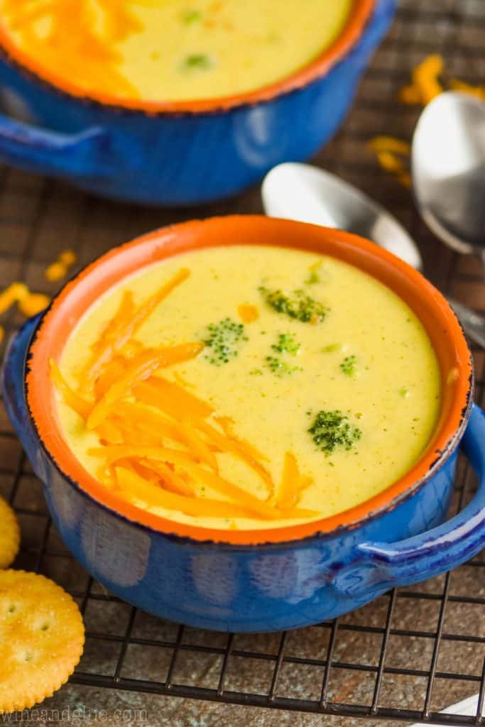 small blue ceramic soup bowl full of broccoli cheddar soup garnished with more cheese and round orange crackers next to the bowl