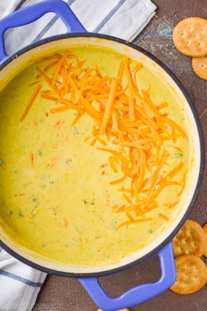 overhead view of a blue dutch oven full of Panera broccoli cheddar soup, garnished with more shredded cheese on a brown surface