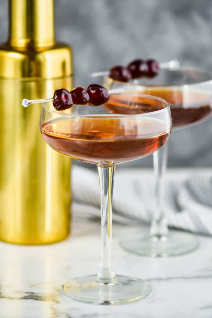 two long champagne coupe glasses filled with a Manhattan drink recipe, garnished with three cherries each, and a gold cocktail shaker in the background