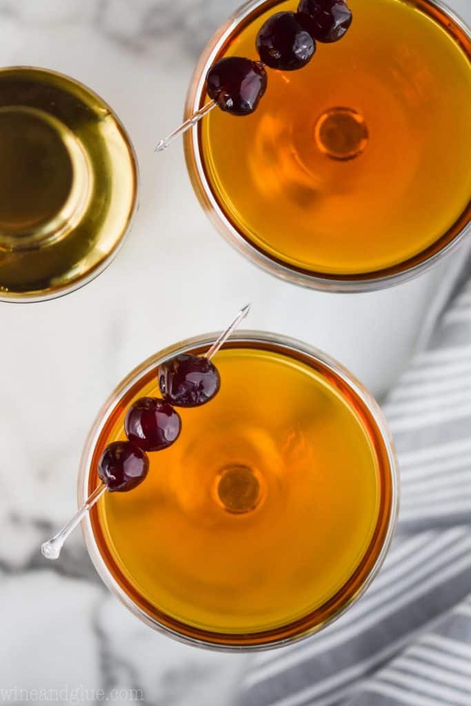 overhead of two Manhattan cocktails in coupe glasses, garnished with three cherries on a plastic toothpick