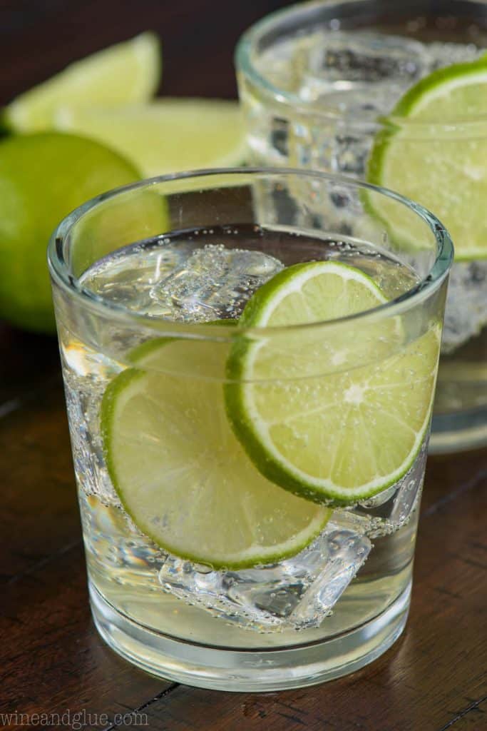 up close view of a gin and tonic recipe in a small tumbler with two lime slices and ice and another glass in the background