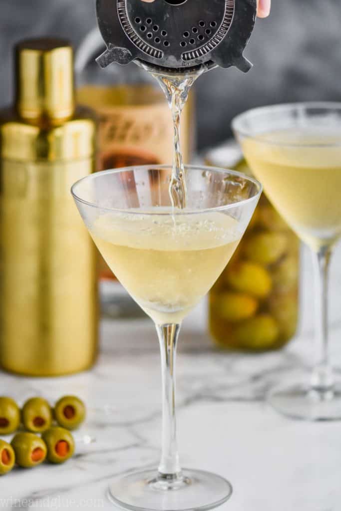 dirty martini recipe being poured through a strainer into a martini glasss with another martini in the background and a gold cocktail shaker