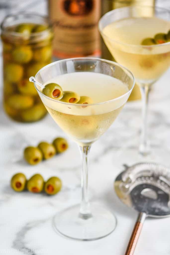 a martini glass full of vodka martini dirty, jar of olives in the background, and strainer next to it