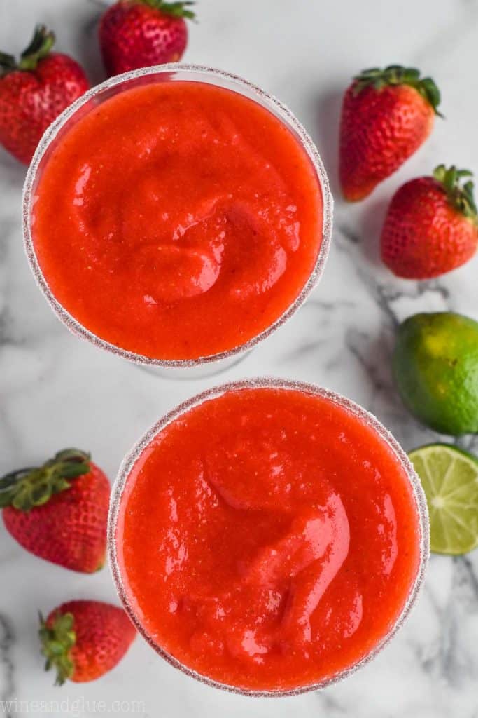 overhead view of two classic margarita glasses rimmed with sugar and filled with red strawberry margaritas, you can see fresh strawberries and limes beneath them