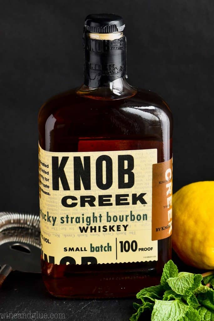 a bottle of knob creek whiskey surrounded by a cocktail strainer, fresh mint, and a lemon