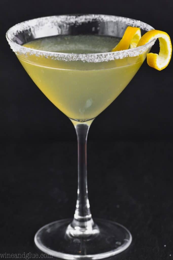 martini glass on a black background rimmed with sugar, full of a lemon drop martini and garnished with a lemon curl